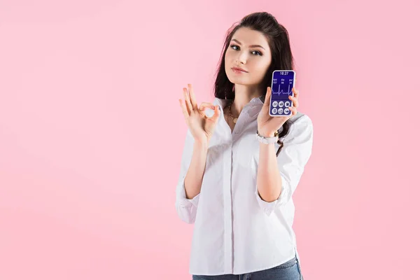 Attractive brunette girl showing ok sign and smartphone with health data on screen, isolated on pink — Stock Photo