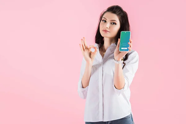 Hermosa chica morena mostrando signo de ok y teléfono inteligente con aplicación de twitter en la pantalla, aislado en rosa - foto de stock