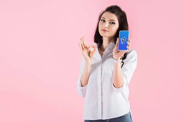 Привабливі брюнетка жінка, показуючи ОК знак і смартфон з shazam app на екрані, ізольовані на рожевий — стокове фото