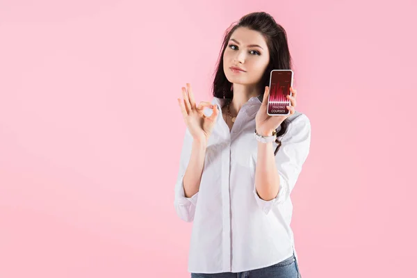 Attractive brunette girl showing ok sign and smartphone with trading courses on screen, isolated on pink — Stock Photo