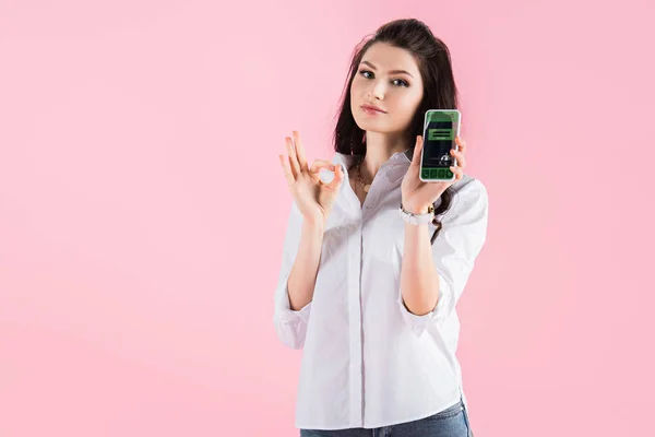 Attractive brunette girl showing ok sign and smartphone with booking app on screen, isolated on pink — Stock Photo