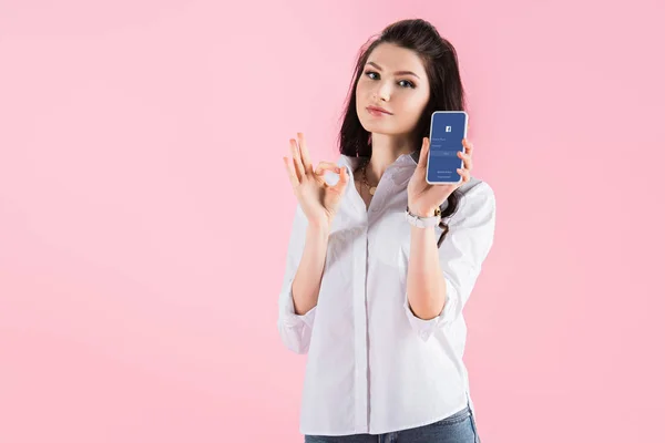 Atractiva mujer joven que muestra signo de ok y teléfono inteligente con aplicación de Facebook en la pantalla, aislado en rosa - foto de stock
