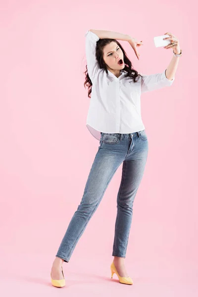 Brunette girl grimacing, gesturing and taking selfie on smartphone, isolated on pink — Stock Photo