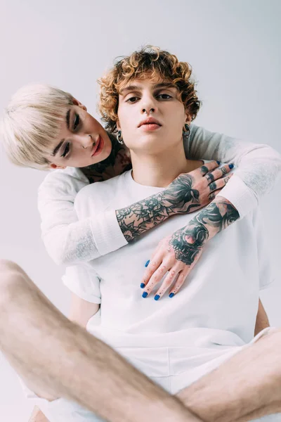 Блондинка з татуюваннями обіймає хлопця з кучерявим волоссям ізольовано на сірому — стокове фото