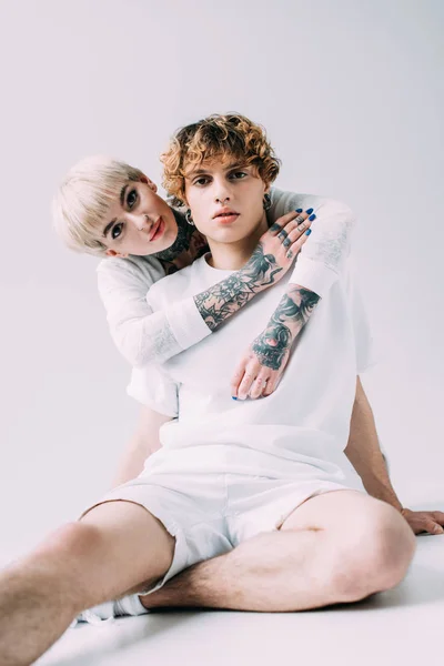 Rubia mujer con tatuajes abrazando novio con pelo rizado aislado en gris - foto de stock