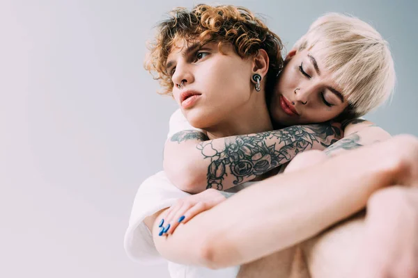 Блондинка з татуюваннями обіймає чоловіка з кучерявим волоссям ізольовано на сірому — стокове фото