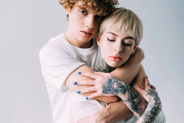 Guapo novio con pelo rizado abrazando mujer rubia con tatuajes aislados en gris - foto de stock