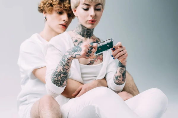 Selective focus of blonde tattooed woman holding audio tape while sitting with boyfriend isolated on grey — Stock Photo