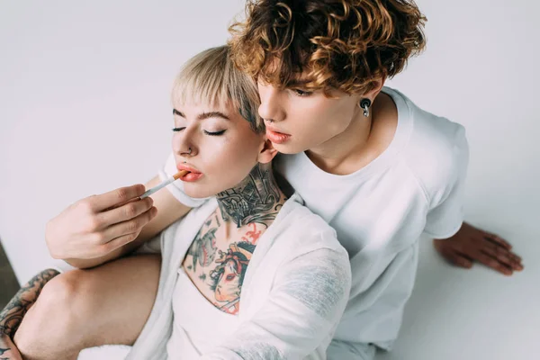 Hombre guapo con el pelo rizado poner cigarrillo en la boca de rubia chica tatuada aislado en gris - foto de stock