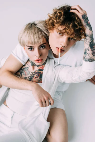 Handsome man with curly hair holding cigarette in mouth while sitting with blonde tattooed girl isolated on grey — Stock Photo
