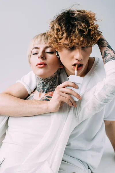 Mujer tatuada abrazando hombre guapo con pelo rizado sosteniendo cigarrillo en la boca aislado en gris - foto de stock