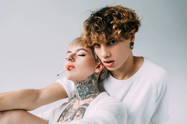 Tattooed woman holding cigarette in mouth near handsome man with curly hair isolated on grey — Stock Photo
