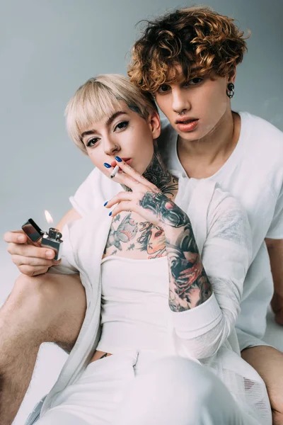 Blonde tattooed girl holding cigarette in mouth near handsome man with lighter in hand isolated on grey — Stock Photo