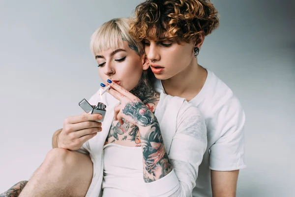 Блондинка татуйована дівчина палить цигарку біля красивого чоловіка з запальничкою в руці ізольовано на сірому — стокове фото