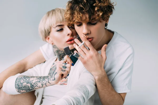 Блондинка татуйована дівчина тримає запальничку біля чоловіка з цигаркою в роті ізольована на сірому — стокове фото