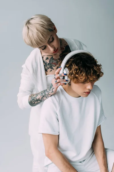 Блондинка татуйована дівчина тримає навушники біля чоловіка з кучерявим волоссям слухаючи музику ізольовано на сірому — стокове фото
