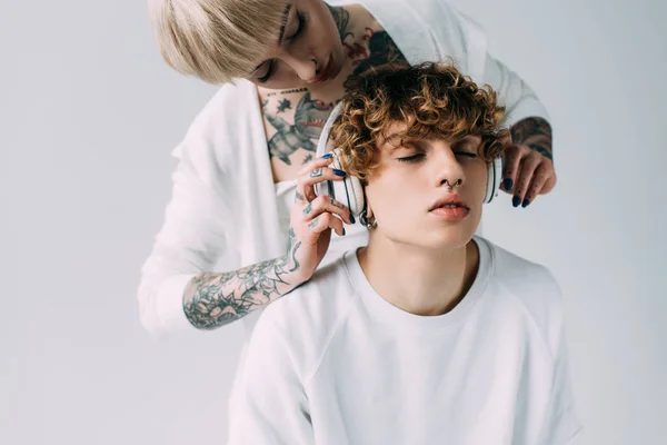 Menina atraente com tatuagens segurando fones de ouvido perto do homem com olhos fechados ouvir música isolada no cinza — Fotografia de Stock