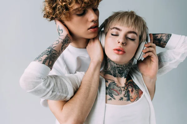 Bell'uomo che tiene le cuffie della ragazza tatuata ad occhi chiusi ascoltando musica isolata su grigio — Foto stock