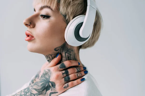 Блондинка с татуировками слушает музыку в наушниках, изолированных на сером — стоковое фото