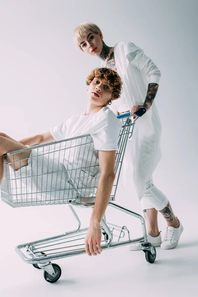 Atraente loira namorada empurrando carrinho de compras com namorado dentro isolado no cinza — Fotografia de Stock