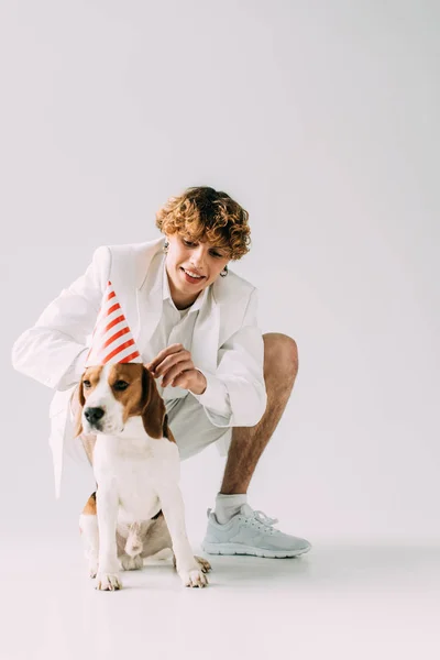 Homme heureux avec des cheveux bouclés portant casquette de fête sur chien beagle sur fond gris — Photo de stock