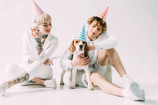 Cheerful couple wearing party caps sitting with beagle dog on grey background — Stock Photo