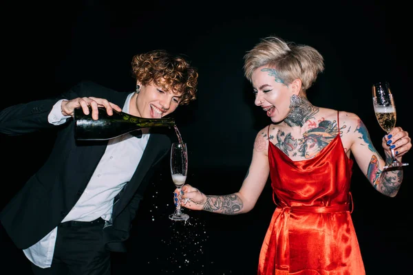 Man with curly hair pouring champagne into glass of cheerful woman in red dress isolated on black — Stock Photo