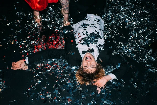 Homem alegre deitado no chão com confete e rindo perto da namorada isolada no preto — Fotografia de Stock