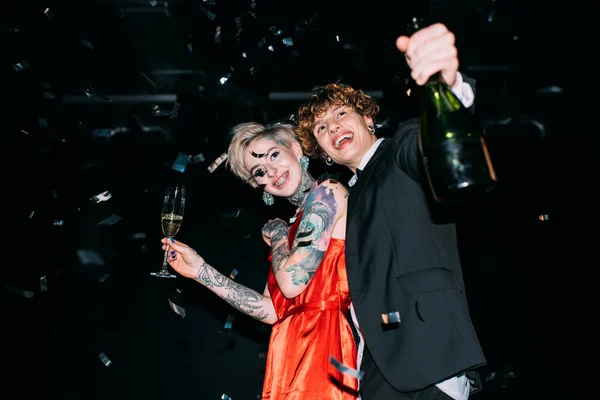 Mulher feliz em vestido vermelho segurando copo de champanhe perto de namorado com garrafa e sorrindo no fundo preto — Fotografia de Stock