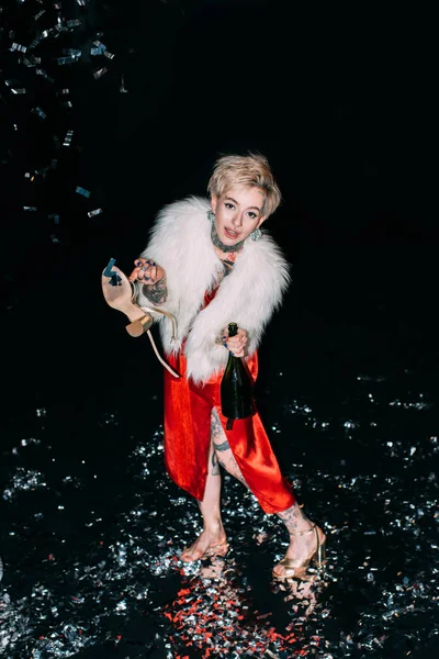 Blonde woman with tattoos holding bottle of champagne and shoe isolated on black — Stock Photo