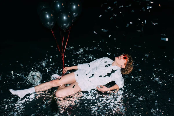 Bel homme aux lunettes de soleil couché sur le sol et tenant des ballons isolés sur noir — Photo de stock