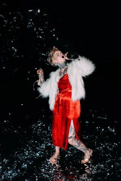 Blonde woman with tattoos drinking alcohol from bottle isolated on black — Stock Photo