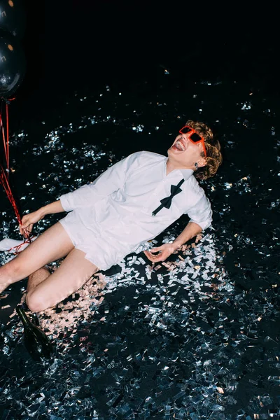 Cheerful man lying on floor with confetti and smiling isolated on black — Stock Photo