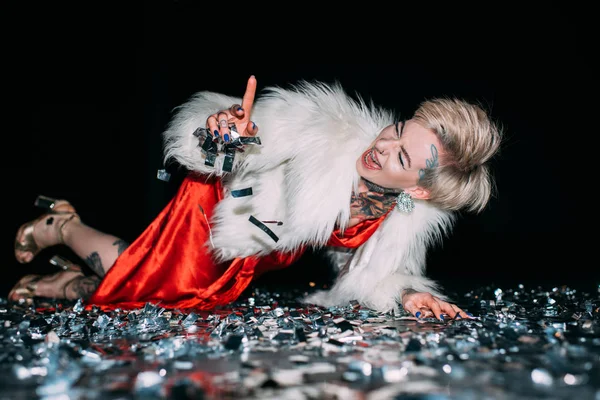 Cheerful blonde woman pointing with finger while lying in floor with confetti isolated on black — Stock Photo