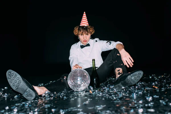 Homme aux cheveux bouclés assis dans la casquette de fête avec boule disco isolé sur noir — Photo de stock