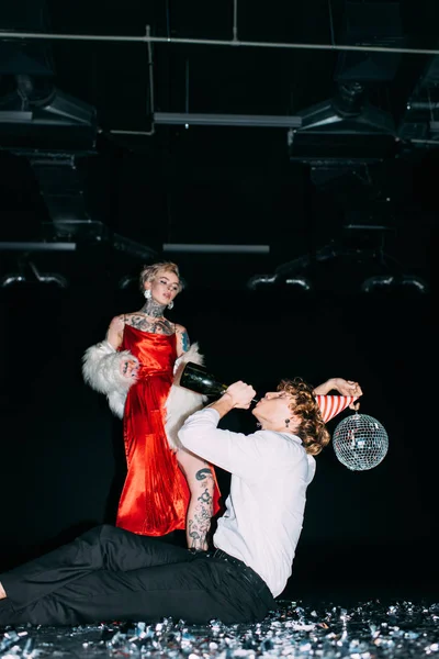 Blonde woman looking at man holding disco ball and drinking from bottle on black background — Stock Photo