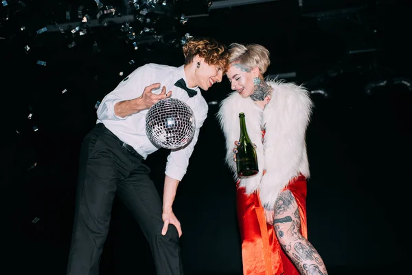 Homme tenant boule disco et debout près de la femme avec bouteille de boisson alcoolisée sur le parti sur fond noir — Photo de stock