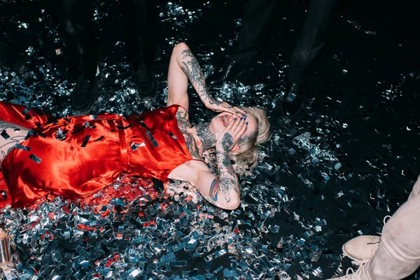 Drunk woman covering face while lying on floor with confetti near people on party isolated on black — Stock Photo