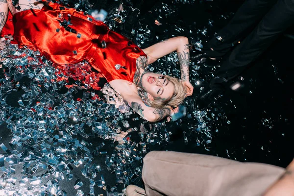 Ivre femme couché sur le sol avec confettis près de personnes sur partie isolé sur noir — Photo de stock