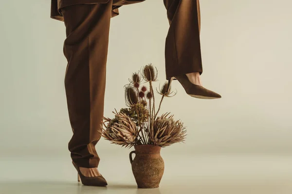 Vista de sección baja de la mujer elegante posando en jarra de arcilla con flores secas en beige - foto de stock