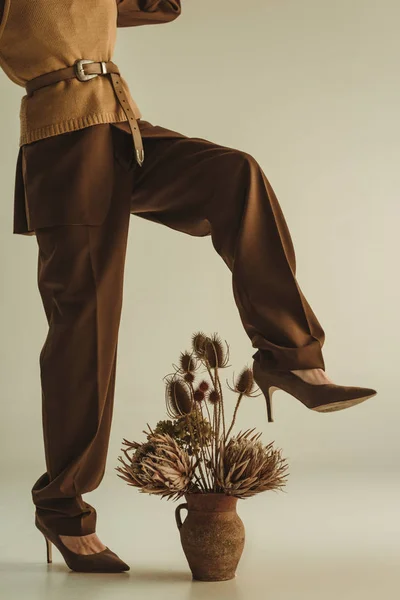 Vista parziale di ragazza alla moda in posa a brocca di argilla con mazzo di fiori secchi su beige — Foto stock