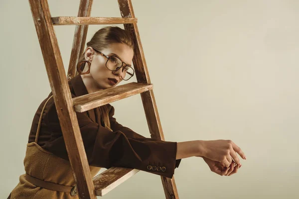 Mujer joven de moda en estilo retro posando cerca de escalera de madera aislada en beige - foto de stock