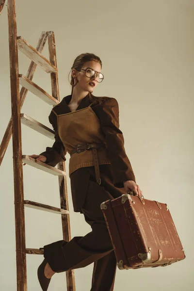 Attractive woman posing with vintage suitcase near ladder isolated on beige — Stock Photo