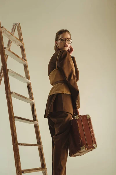 Красивая стильная девушка позирует с винтажным мешок для путешествий рядом с лестницей изолированы на бежевый — стоковое фото