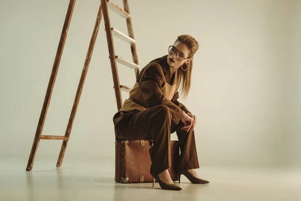 Красивая стильная женщина сидит на винтажном чемодане возле лестницы на бежевом — стоковое фото