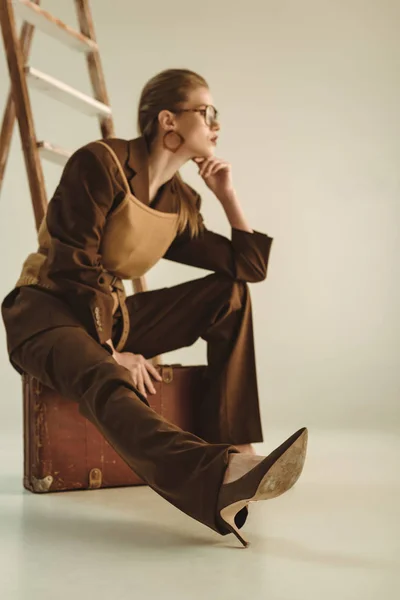 Enfoque selectivo de chica elegante sentado en la maleta vintage cerca de escalera en beige - foto de stock
