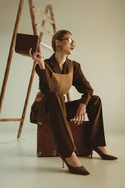 Menina atraente em estilo vintage segurando livro e sentado perto de escada em bege — Fotografia de Stock