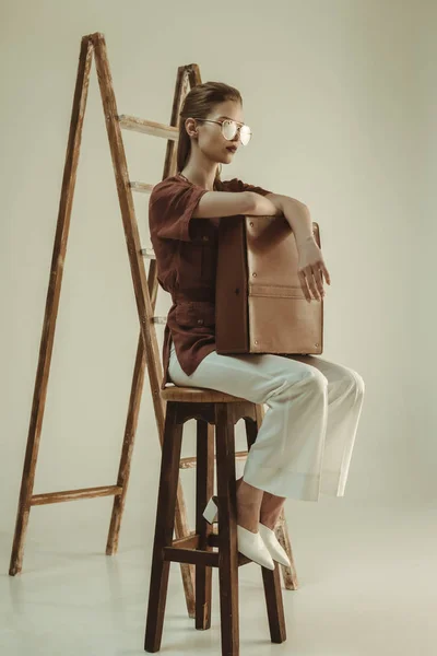 Attraktive junge Frau hält Ledertasche in der Hand und sitzt neben Leiter auf beige — Stockfoto