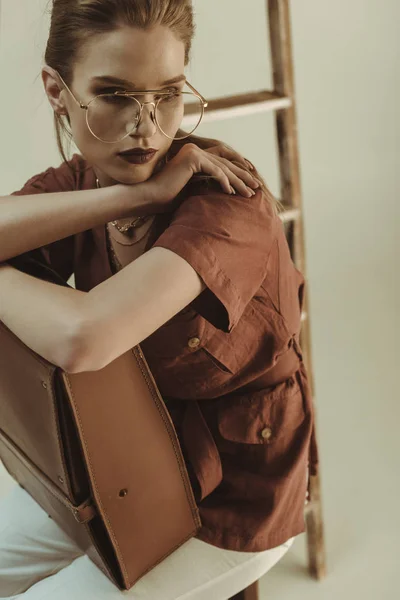 Красива молода жінка тримає шкіряний мішок і сидить біля сходів ізольовано на бежевому — стокове фото