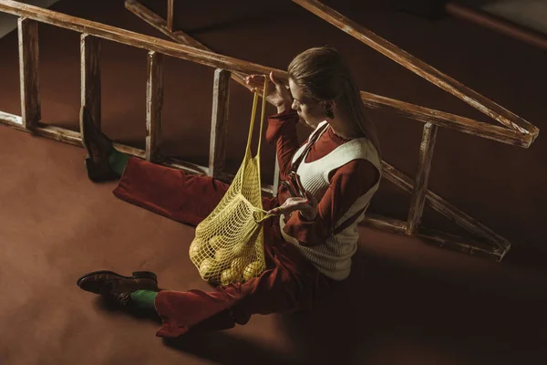 Attractive girl with lemons in string bag posing near ladder — Stock Photo